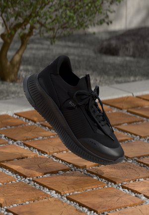 Baskets noires Boss avec lacets sur des carreaux carrés en bois en extérieur près d’un petit arbre et d’un sol en pierre.