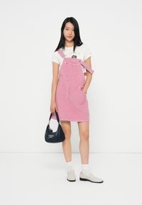 Rosa Cord-Wolloverallkleid mit verstellbaren Trägern, Fronttaschen und lockerer Passform, kombiniert mit einem Grafik-T-Shirt und grauen Slippern.