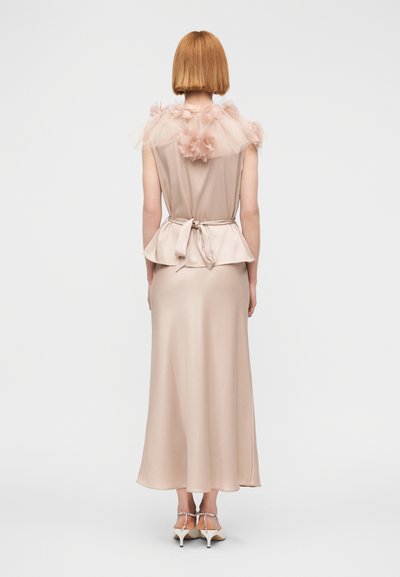 Femme aux cheveux roux coupés au carré, portant une robe beige sans manches avec du tulle floral sur les épaules et une ceinture nouée à la taille, de dos.