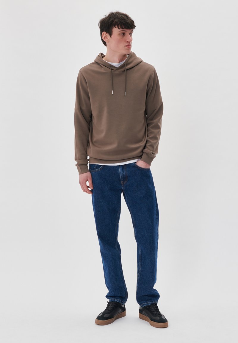 Joven de pie con una mano en el bolsillo, vestido con sudadera marrón, jeans azules y zapatillas negras, mirando de lado contra un fondo liso.