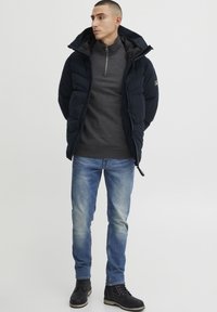 Marineblå pufjakke med hætte, grå lynlåsgenser, lyse Wasch-jeans og sorte snørebåndsstøvler. Glatte teksturer og minimalt hardwaredetaljer.