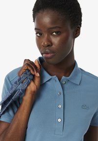 Vaaleansininen polo-paita teksturoidusta kankaasta, kolmella napilla varustettu helma ja brodattu logo. Malli pitää kädessään kuviollista hihnaa.