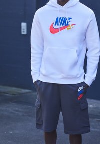 Vitt Nike-hoodie med flerfärgad logotyp, tillsammans med grå cargoshorts som har flera Nike-logotyper i röd, blå och vit. Avslappnad sportig klädsel.