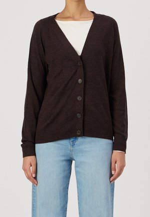 Kvinde iført en mørkebrun cardigan med knapper over en hvid skjorte og lyseblå jeans, stående mod en ensfarvet baggrund.