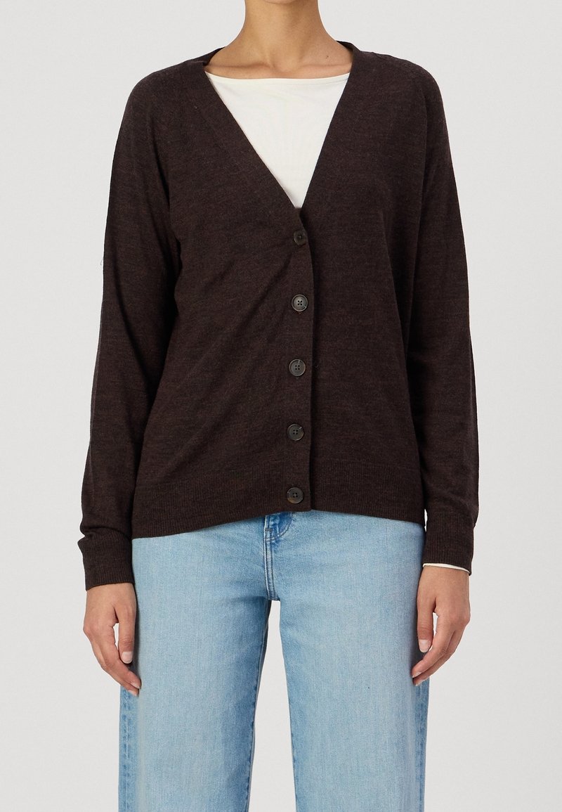 Femme portant un cardigan marron foncé à boutons sur une chemise blanche avec un jean bleu clair, debout devant un fond uni.