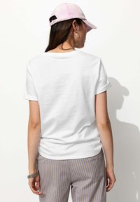 T-shirt en coton blanc avec des manches retroussées, associé à un pantalon rayé gris et jaune. Une casquette rose clair est portée, vue de dos.