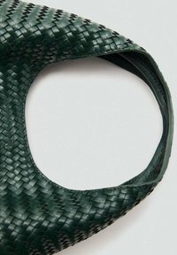 Bolso de mano verde tejido con un diseño abierto en la parte superior, que presenta un borde de cuero suave y un patrón texturizado en todo el cuerpo.