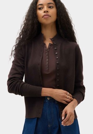 Frau mit langen lockigen Haaren trägt eine dunkelbraune, geknöpfte Strickjacke über einem passenden Henley-Shirt und blauen, hoch taillierten Jeans und richtet ihren Ärmel.