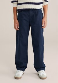 Námornícky modré nohavice s širokými nohavicami vyrobené z bavlnenej zmesi, s navliekaným dizajnom a bočnými vreckami. Nosia sa s bielymi teniskami.