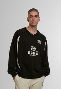 Juodas megztinis su baltais akcentais, su V formos iškirpte, ilgomis rankovėmis ir spausdintu logotipu. Minkšta tekstūra; laisvės pojūtį suteikiantis dizainas.
