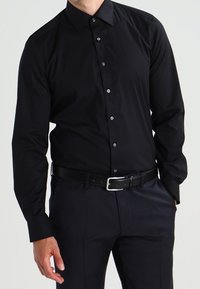 Camicia da uomo nera a maniche lunghe in tessuto satinato con colletto classico, bottoni frontali e vestibilità slim, abbinata a pantaloni scuri e una cintura.