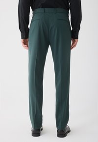 Mężczyzna ubrany w ciemnozielone dopasowane spodnie, czarną koszulę schowaną w spodniach oraz czarne buty eleganckie, stojący z rękami swobodnie opuszczonymi wzdłuż ciała.