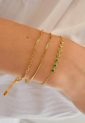 Drei zarte Goldarmbänder an einem Handgelenk, eines mit Gliederkette, eines glatt und eines mit grünen Edelsteinen verziert.