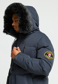 Jūrinės spalvos pūkinė striukė su kailiniu gobtuvu, turinti priekinį užtrauktuką, dvi išorines kišenes ir dekoratyvinę siuvinėtą emblema ant rankovės.