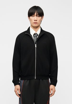 Ung mand med glat sort hår iført en sort lynlåstrøje, hvid skjorte med krave, sort slips og sorte bukser med røde striber.