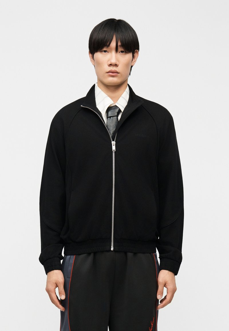 Νέος άνδρας με ίσια μαύρα μαλλιά που φοράει μαύρο μπουφάν με φερμουάρ, λευκό πουκάμισο με γιακά, μαύρη γραβάτα και μαύρο παντελόνι με κόκκινες ρίγες.