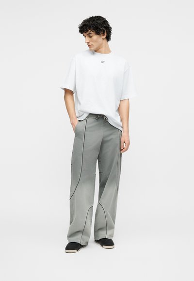 Νεαρός με σγουρά μαλλιά, φορεί λευκό ευρύχωρο T-shirt και χαλαρά γκρι παντελόνια με μαύρη επένδυση, στέκεται με τα χέρια στις τσέπες.