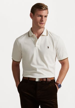 Maglietta polo bianca con bordo del colletto marrone, caratterizzata da un piccolo logo sul petto. Indossata con pantaloni in corduroy marroni e una cintura abbinata.