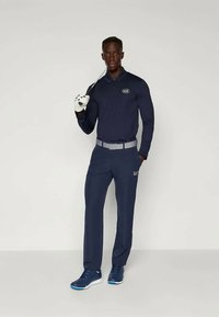 Námořné golfové polo s dlouhými rukávy a logem, spárované s odpovídajícími kalhotami a modrými sportovními botami. Model drží golfovou hůl a rukavici.