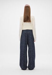 Suéter de malha creme combinado com calças largas azuis marinho, apresentando um tecido texturizado e um design pregueado. Vista pela parte de trás.