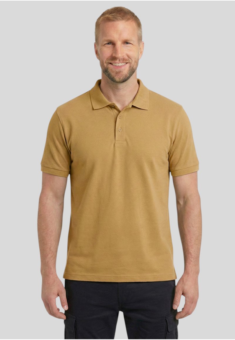 Homme souriant aux cheveux courts et à la barbe portant un polo jaune moutarde et un pantalon cargo noir, debout devant un fond gris uni.