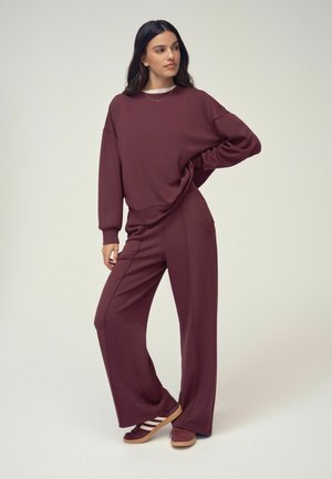 Kvinde stående med hånd på hoften iført en bordeaux sweatshirt og vidde ben bukser med matchende sko mod en ensfarvet baggrund.