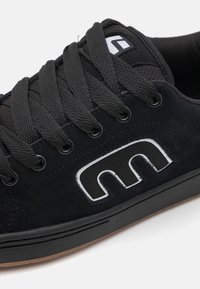 Black suede sneaker with textured surface, flat sole, and black laces. Features a white logo accent on the side. Classic design.