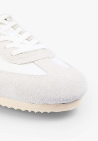 Zapatilla de ante y sintético en gris claro y blanco, con punta redondeada, diseño de cordones y suela texturizada para agarre.
