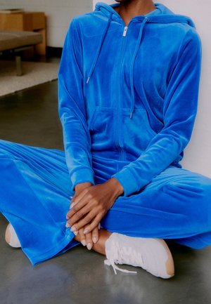 Personne assise en tailleur sur le sol, portant un sweat à capuche zippé bleu vif et un pantalon assorti avec des baskets blanches, mains reposant sur les chevilles.