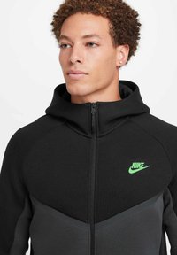 Sudadera con cremallera negra y logo de Nike verde. Presenta un panel gris en contraste en el pecho y una capucha con cordón. Tejido texturizado.