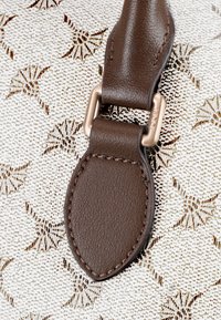 Brun læderrem fastgjort til en hvid tekstureret baggrund med et diskret stjernemønster. Hardware med en lys metallisk finish.