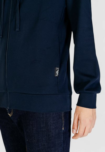 Felpa con zip color navy realizzata in tessuto testurizzato, con un'etichetta logo discreta sul lato e polsini elasticizzati. Indossata con jeans di denim scuri.