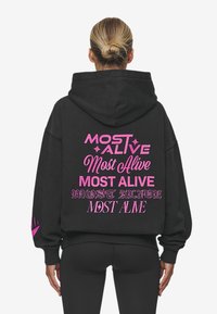 Černá mikina s kapucí, na které je vzadu výrazný růžový nápis "MOST ALIVE" napsaný různými fonty. Vyrobena z měkké látky, má klasickou kapuci a uvolněný střih.