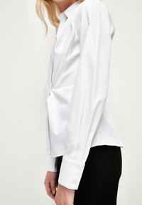 Chemise blanche à boutons avec manches longues, silhouette ajustée et ourlet incurvé. Fabriquée en tissu lisse, léger, avec une texture subtile.