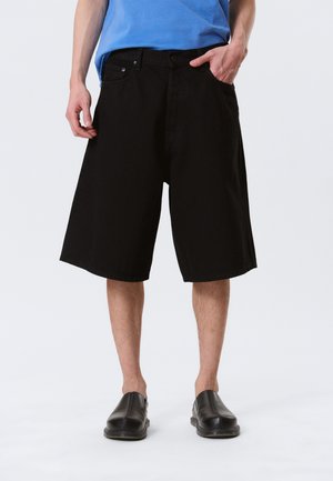 Personne portant un short noir ample jusqu'aux genoux, une chemise bleue et des chaussures noires à enfiler, debout avec une main dans la poche.