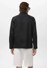 Sort jakke med et struktureret design, lange ærmer og Krave. Båret over lyse shorts. Visningen viser detaljerne bagpå.