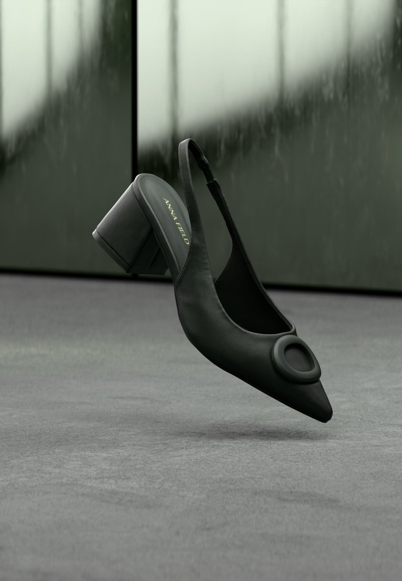 Μαύρη δερμάτινη γόβα με λουράκι στο πίσω μέρος, ψηλό τακούνι, μυτερή μύτη και λεπτομέρεια με κυκλικό αγκράφα, τοποθετημένη σε γκρι δάπεδο με θολό φόντο.