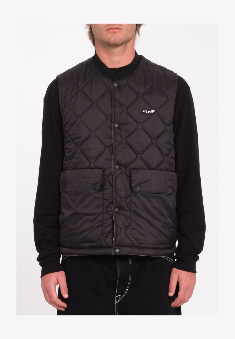 Gilet rembourré noir avec un design matelassé, comportant deux poches avant, des boutons-pression et un petit logo sur la poitrine. Porté par-dessus une chemise à manches longues.