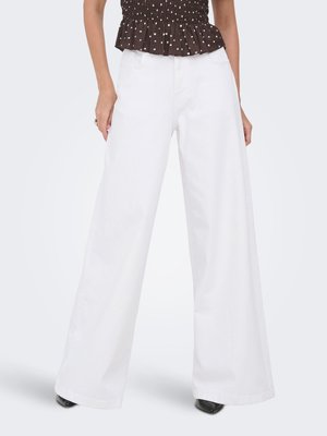Femme portant un pantalon blanc taille haute à jambes larges, associé à un haut noir à pois et des chaussures noires pointues, debout les mains le long du corps.