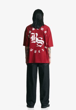 Rød bomulds oversized t-shirt med stort hvidt grafiktryk på bagsiden, parret med mørke denimbukser og sorte sko.