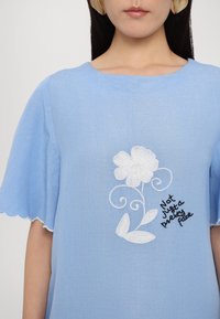Helles blaues kurzärmliges Blusenshirt mit weißer Blumenstickerei und dem Text "Nicht nur ein hübsches Gesicht" in Schwarz, mit gewellten Saumakzenten.
