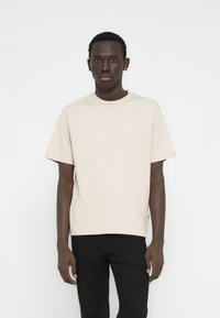 Ανοιχτό μπεζ βαμβακερό μπλουζάκι με λαιμόκοψη γύρω από το λαιμό, κοντά μανίκια και μικρή λευκή επιγραφή μπροστά, συνδυασμένο με μαύρο παντελόνι.