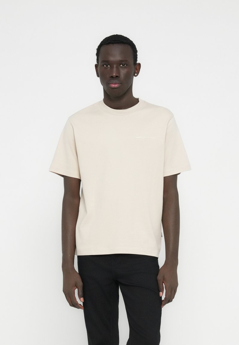 Ανοιχτό μπεζ βαμβακερό μπλουζάκι με λαιμόκοψη γύρω από το λαιμό, κοντά μανίκια και μικρή λευκή επιγραφή μπροστά, συνδυασμένο με μαύρο παντελόνι.