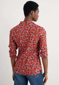 Camicia a maniche lunghe con bottoni rossi, caratterizzata da un tessuto testurizzato, un motivo floreale in vari colori e un orlo curvo.