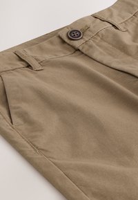Close-up of beige pants waistband with button closure, belt loops, and topstitching details on fabric.