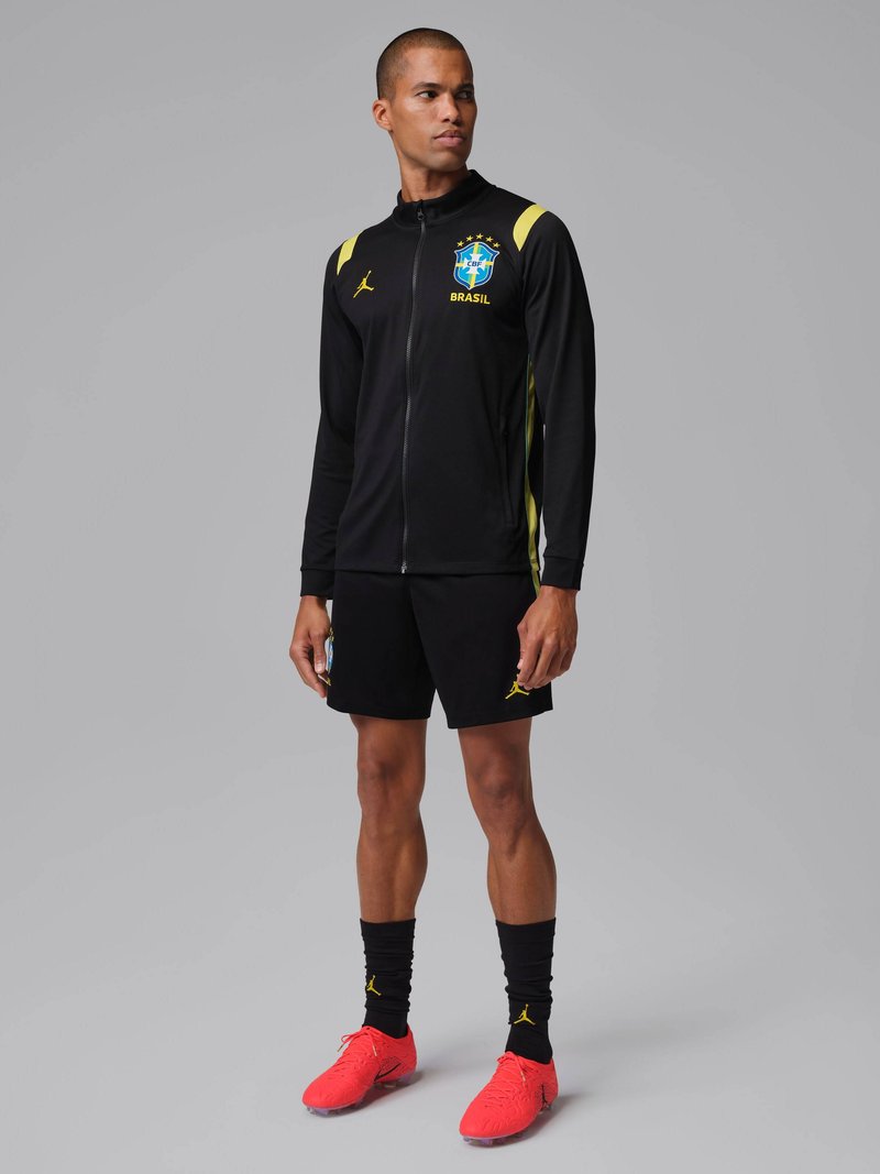Hombre con chaqueta de entrenamiento de fútbol de Brasil negra con detalles amarillos, pantalones cortos negros, calcetas negras y tacos rojos brillantes, de pie contra un fondo gris.