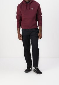 Felpa marrone con cappuccio, tasca frontale e logo, abbinata a pantaloni neri e sneakers nere con strisce bianche. Tessuto testurizzato, vestibilità casual.