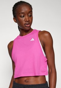Rosa, ärmelloses Top in verkürzter Form aus glattem Material mit weißem Logoakzent. Es hat einen lockeren Schnitt und einen runden Halsausschnitt; kombiniert mit dunklen Shorts.