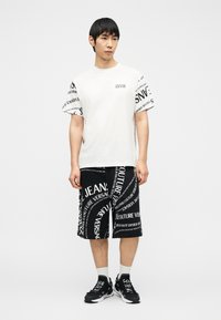 Άνδρας που φοράει λευκό T-shirt και μαύρο σορτς με μεγάλα λευκά τυπωμένα λογότυπα, συνδυασμένα με μαύρα και λευκά αθλητικά παπούτσια και λευκές κάλτσες.