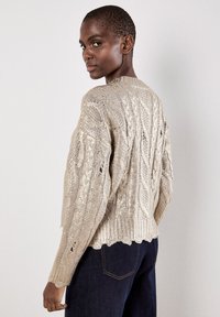 Maglione lavorato a maglia, beige, con un design a trecce texturizzate, orlo scallop e maniche lunghe. Indossato con jeans blu scuro, che mostrano una vestibilità rilassata.